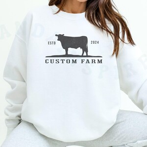 Puede incluir: Sudadera blanca con una silueta negra de una vaca y el texto "ESTO 2024 CUSTOM FARM".