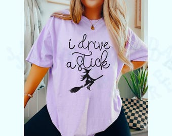 May include: A light purple t-shirt with the phrase "i drive a stick" in black script, along with a silhouette of a witch on a broom. The shirt is a short-sleeved, oversized fit.