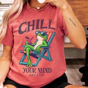 May include: Red-orange t-shirt with a graphic of a frog in sunglasses relaxing in a beach chair, holding a drink. The text "CHILL YOUR MIND SUMMER TIME" is above and below the frog. The shirt is a casual summer wear item.