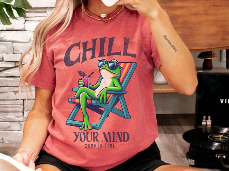 May include: Red-orange t-shirt with a graphic of a frog in sunglasses relaxing in a beach chair, holding a drink. The text "CHILL YOUR MIND SUMMER TIME" is above and below the frog. The shirt is a casual summer wear item.