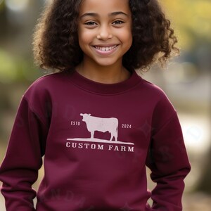 Puede incluir: Una sudadera color burdeos con una silueta blanca de una vaca y el texto "ESTD 2024 CUSTOM FARM".