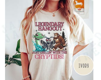 May include: Ivory t-shirt with a graphic print of a green sea serpent, a moth, and a Bigfoot. The text on the shirt reads "Legendary Hangout Just Chillin' With The Cryptids!"