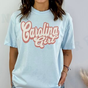 May include: A light blue t-shirt with the text "Carolina Girl" in a retro-style font. The text is in pink and white.