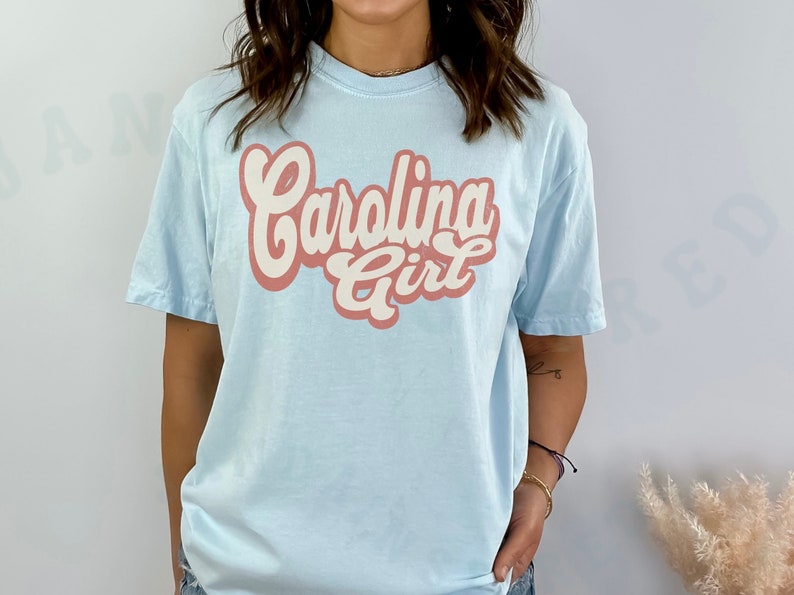 May include: A light blue t-shirt with the text "Carolina Girl" in a retro-style font. The text is in pink and white.