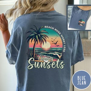 May include: A blue jean t-shirt with a graphic of a palm tree, sunset, and waves. The text "Beach More Worry Less" is written in a circle around the graphic. The text "Chasing Sunsets" is written below the graphic.