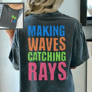 May include: A dark gray t-shirt with the text "MAKING WAVES CATCHING RAYS" printed on the back in colorful letters. The text is in a distressed font style.