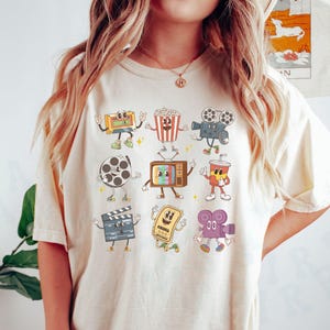 May include: A white t-shirt with a graphic print of cartoon characters representing different movie-related items, including a film reel, popcorn, a clapperboard, a movie ticket, a camera, and a television.