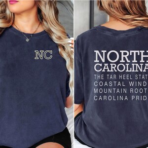 May include: Navy blue t-shirt featuring "NC" with a yellow outline on the front. The back of the shirt displays "NORTH CAROLINA" in white block letters, along with state slogans. A casual, comfortable tee.