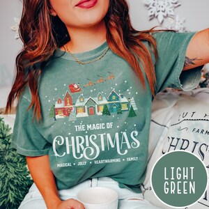 May include: A light green t-shirt with a Christmas-themed design featuring a row of houses with snow on the roofs and the text "The Magic of Christmas" with the words "Magical, Jolly, Heartwarming, Family" underneath.
