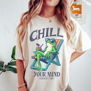 May include: Cream-colored t-shirt featuring a cartoon frog in sunglasses relaxing in a beach chair with a drink. The shirt has the words "CHILL YOUR MIND SUMMER TIME" in a distressed font.