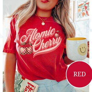 May include: Red t-shirt with a retro graphic design that says "Atomic Cherry" with a checkered cherry illustration.