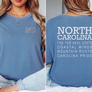 May include: A pale blue t-shirt with the letters "NC" on the front and the words "NORTH CAROLINA" and other text on the back. The shirt has a crew neck and short sleeves. The text on the back includes "THE TAR HEEL STATE", "COASTAL WINDS", "MOUNTAIN ROOTS", and "CAROLINA PRIDE".