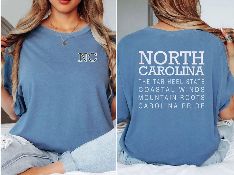 May include: A pale blue t-shirt with the letters "NC" on the front and the words "NORTH CAROLINA" and other text on the back. The shirt has a crew neck and short sleeves. The text on the back includes "THE TAR HEEL STATE", "COASTAL WINDS", "MOUNTAIN ROOTS", and "CAROLINA PRIDE".