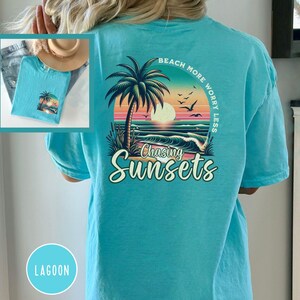 May include: A turquoise blue t-shirt with a graphic print of a palm tree, sunset, and ocean waves. The text "Beach More Worry Less" is printed in a circle around the graphic. The text "Chasing Sunsets" is printed below the graphic.