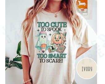 May include: A white t-shirt with the text "Too Cute To Spook Too Smart To Scare!" in green and black lettering. The design features a ghost holding a pencil and a pumpkin holding a pencil. The shirt is available in ivory.