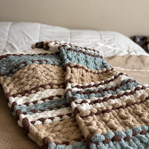 May include: A chunky knit blanket in light blue, brown, cream, and tan. The blanket is folded over a bed, showing its textured, cosy appearance. The handmade blanket features a striped pattern, perfect for adding warmth and style to any bedroom.