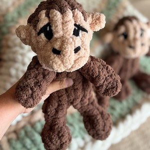 以下が含まれることがあります： ダークブラウンとライトベージュの毛糸で作られた猿のぬいぐるみ。この猿は、質感のあるふわふわとした外観で、目と鼻には黒い刺繍が施されています。頭部はライトベージュで、ダークブラウンの耳と頭頂部が特徴です。