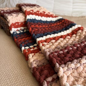 May include: A folded, chunky knit blanket in shades of red, navy, cream, and beige. The blanket has a textured, looped appearance and is folded over on a neutral-colored surface. The blanket appears soft and cozy.
