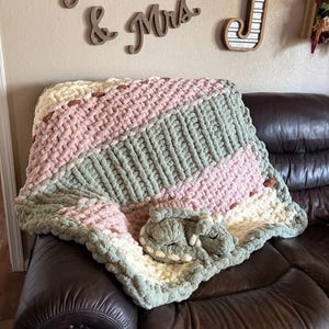 May include: A chunky knit blanket featuring diagonal stripes of cream, dusty pink, and sage green yarn. Resting on top of the blanket is a small, crocheted dragon plush toy in a matching sage green color with cream-colored accents on its back.