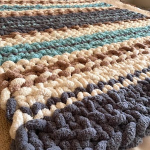 Op de afbeelding: Een grof gebreide plaid met horizontale strepen in de kleuren donkergrijs, crème, lichtbruin en groenblauw. De deken is gemaakt van dikke, zachte garenlussen in een gestructureerd patroon, wat zorgt voor een volumineuze en zachte uitstraling.