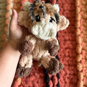 Puede incluir: Un juguete de peluche hecho a mano con forma de vaca, con un hocico color crema, cuerpo marrón claro y pezuñas marrones oscuras. La vaca tiene ojos negros y un aspecto esponjoso y texturizado. El juguete se sostiene contra una manta colorida y texturizada.