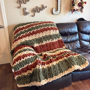 Könnte beinhalten: Eine grob gestrickte Decke in Olivgrün, Creme und Rostrot, drapiert über einem dunkelbraunen Ledersofa. Die Decke hat horizontale Streifen und ein dickes, strukturiertes Gewebe, ideal für gemütliche Dekoration.
