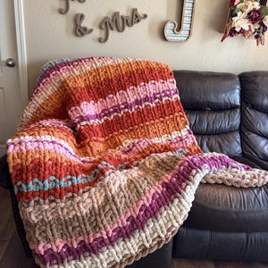 May include: A chunky knit blanket in shades of orange, pink, and beige is draped over a brown leather couch. The blanket features a textured, striped pattern. Decorative wooden letters spell "Mr & Mrs" on the wall.