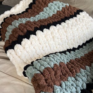 May include: A thick, hand-knitted blanket with a chevron design. The blanket has stripes of white, brown, black, and a soft teal. The close-up shows the soft, plush texture of the yarn.