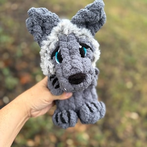 Op de afbeelding: Een handgemaakte gehaakte knuffel in de vorm van een grijze wolf met een lichaam van getextureerd garen, pluizige witte imitatiebont accenten rond het hoofd, grote puntige oren, geborduurde ogen met felblauwe irissen en een kleine zwarte neus.