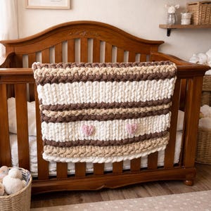 May include: A chunky knit baby blanket in a crib. The blanket features alternating stripes of cream, light brown, and dark brown. Two small pink hearts are placed on the cream stripe. The crib is made of dark wood.