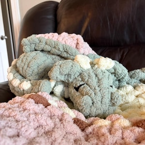 Könnte beinhalten: Ein gehäkeltes Drachen-Stofftier aus dickem, strukturiertem Garn in Salbeigrün, zartem Rosa und Creme. Der Drache hat einen abgerundeten Kopf mit kleinen cremefarbenen Hörnern, geschlossene Augen und einen zusammengerollten Körper.