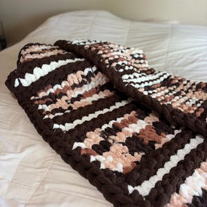 May include: A handmade, chunky knit blanket in shades of brown, white, and blush pink. The blanket features a repeating pattern of horizontal stripes and a thick, braided border. The soft, textured blanket is folded on a white bedspread.