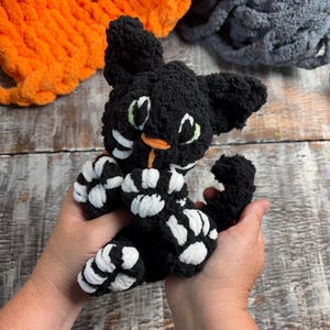 Peut inclure: Un petit chat en peluche fait main, réalisé en fil noir duveteux. Le chat présente des accents en fil blanc sur les pattes et le visage, avec des yeux brodés vert clair et un petit nez orange. Le jouet a des oreilles pointues et une queue recourbée.