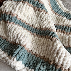 May include: A folded chunky knit blanket with a soft texture. The blanket has a striped pattern with bands of white, muted teal, and light brown. The item appears to be warm and comfortable, ideal for snuggling.