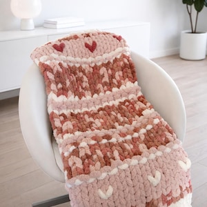 Pode incluir: Um cobertor de malha grossa em tons de rosa, branco e castanho, adornado com detalhes em forma de coração. O cobertor está sobre uma cadeira branca, mostrando seu design texturizado e apelo aconchegante. Um item decorativo para a casa.