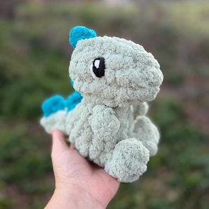 May include: A handmade, light green crochet dinosaur plushie with blue accents. The dinosaur has a textured, fluffy appearance, black and white eyes, and is held in a hand. The plushie is a soft toy.