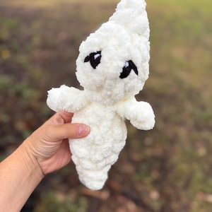 May include: A white crocheted ghost plush toy with a soft, textured yarn finish. The ghost features a pointed top, two small arms extending to the sides, and embroidered black eyes with small white accents, creating a simple, rounded facial expression.