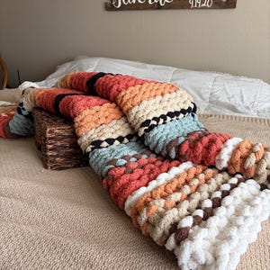May include: A chunky knit blanket in a variety of colors, including coral, orange, cream, and black, is draped over a bed. The blanket is made of thick yarn and has a textured appearance. A wooden sign with the text "But Aug is my Favorite" is visible in the background.