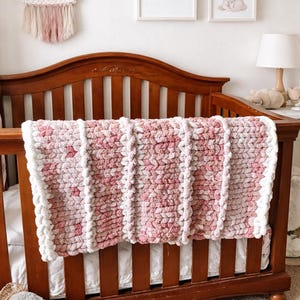 Può includere: Una coperta all'uncinetto rosa e bianca fatta a mano su una culla di legno. La coperta presenta un design strutturato con strisce bianche verticali. La culla si trova in una cameretta con opere d'arte incorniciate e un arazzo in macramè.