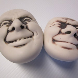 May include: Two white ceramic sculptures of faces with exaggerated expressions. The faces are smiling with their mouths open and their eyes closed. The sculptures are made of a smooth, white ceramic and have a brown glaze on the features.
