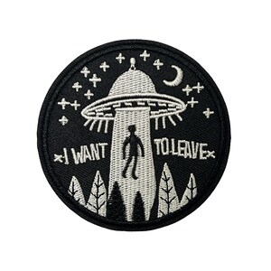 以下が含まれることがあります： 白いUFOのデザイン、シルエット、木、星、そして「I WANT TO LEAVE」というテキストが特徴の、黒い刺繍パッチ。パッチは丸く、黒い縁取りがあります。
