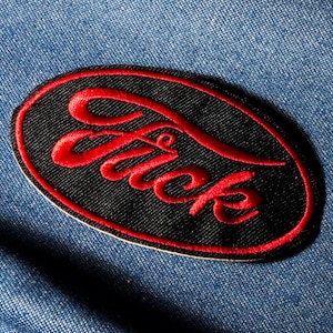 Peut inclure: Un écusson brodé avec le mot "Fuck" en lettres rouges sur un ovale noir. L'écusson a une bordure rouge et est posé sur un fond en denim bleu.