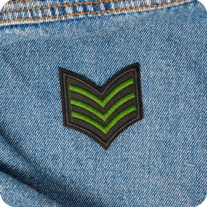 May include: Close-up of a blue denim fabric with a black and green chevron patch. The patch has a black border and three green stripes. The denim has visible stitching at the top.