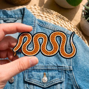 May include: A detailed patch featuring a stylized snake design. The snake is rendered in shades of orange and brown, with a textured pattern, outlined in white and black. The patch is held against a denim jacket.