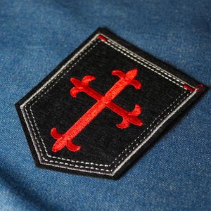 Könnte beinhalten: Schwarzer, gestickter Aufnäher mit einem roten Kreuz-Design. Der Aufnäher hat einen weiß genähten Rand und ist vor einem blauen Jeans-Hintergrund platziert. Das Kreuz ist ein stilisiertes, dekoratives Element.