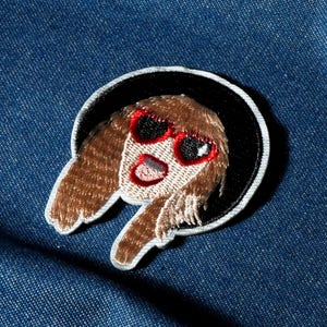 Op de afbeelding: Geborduurde patch met een cartoongezicht met bruin haar en een rode zonnebril, uitkomend uit een zwarte cirkel met een witte rand. Op een blauwe denim achtergrond.