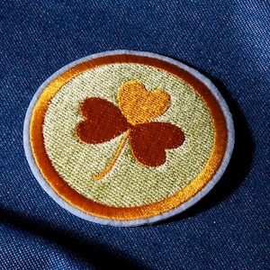 May include: Embroidered shamrock patch. The shamrock is in orange and yellow hues, set against a light green background. The patch has a gold border and a light blue outer edge, on a blue denim background.
