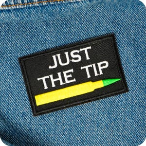 Op de afbeelding: Zwart rechthoekig embleem met de tekst "JUST THE TIP" in het wit. Een gele en groene markeerstift-afbeelding staat onderaan. Het embleem is op een blauwe denim stof genaaid.
