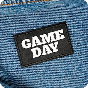 Op de afbeelding: Een zwart rechthoekig embleem met de woorden "GAME DAY" in dikke witte letters is op een blauwe denim stof genaaid. De patch heeft een zwarte rand en is gecentreerd op de denim.
