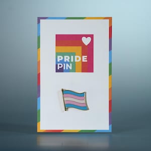 May include: A Pride Pin featuring a transgender flag design. The pin is presented on a card with a rainbow border and a colourful square with a heart and the words "PRIDE PIN". The pin has light blue, pink, and white stripes.